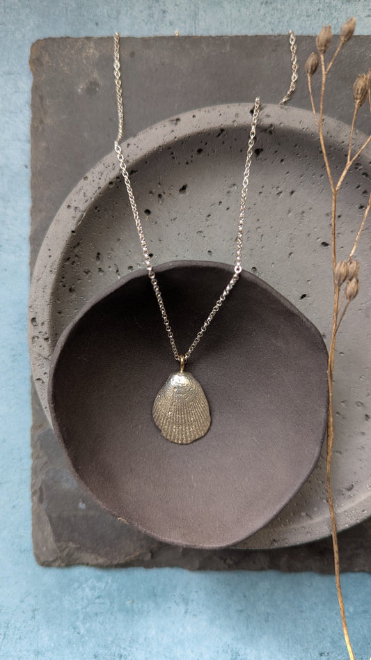 A textured silver pendant necklace shaped like a cockle shell, hanging on a delicate silver chain. The necklace is styled in a flat-lay arrangement over a grey ceramic dish, a square slate tile, and dried grass stems against a light blue background.
